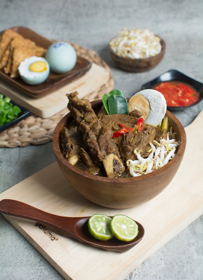 Delicious Indonesian Rawon soup with beef, egg, and sprouts in a wooden bowl, served with sambal and lime.