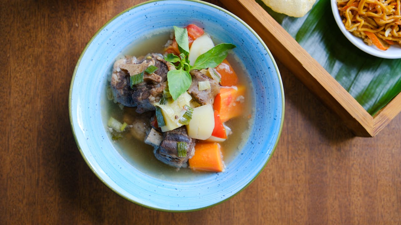 Top view of traditional Indonesian oxtail soup with vegetables, served with side dishes in a vibrant setup.